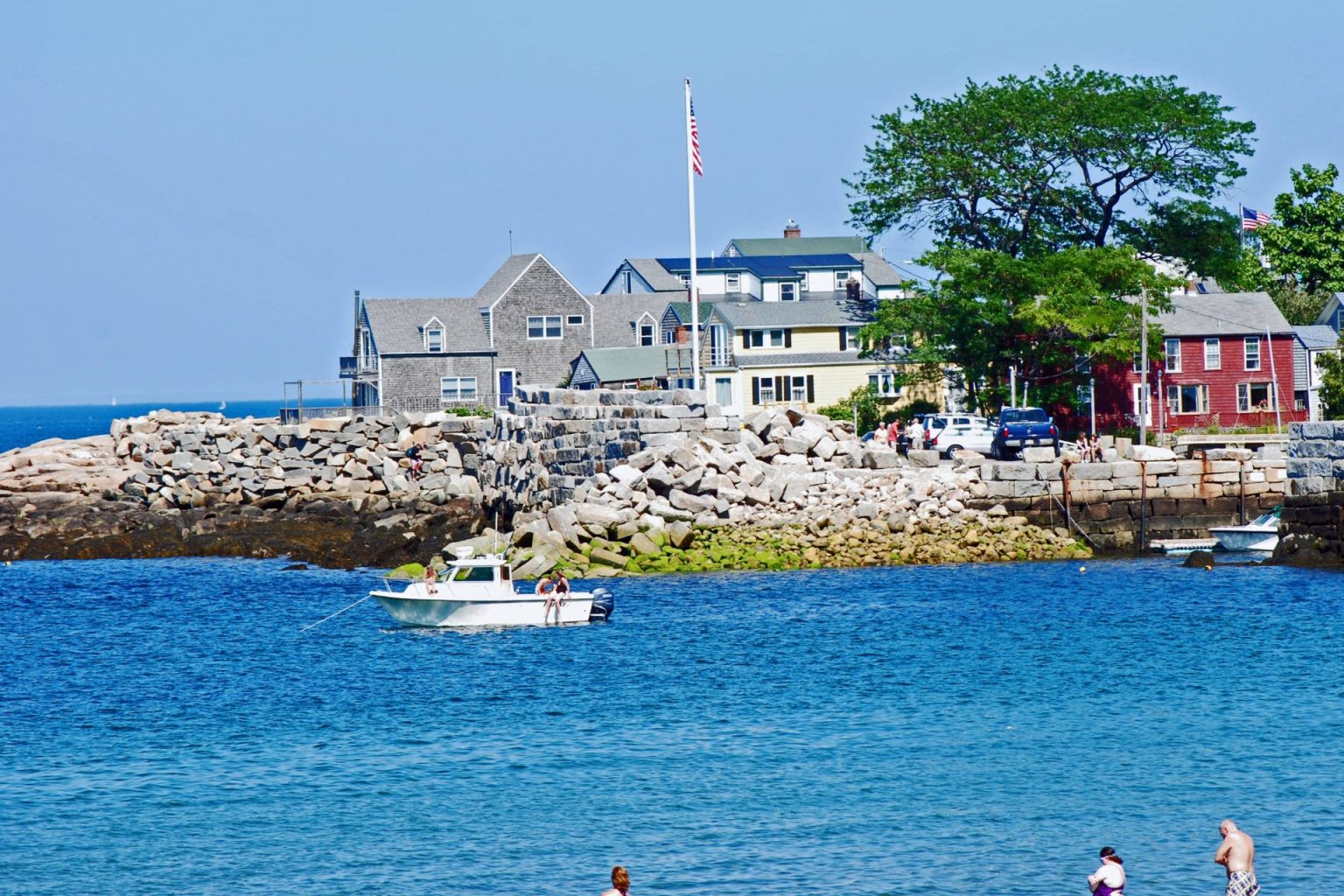 A view of a point on Cape Ann taht has docks, a flag pole, and various New England styled building perched on top of a rock wall