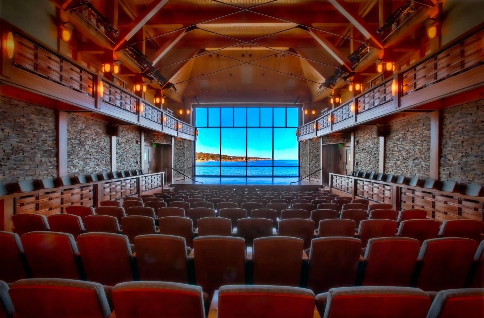 Behind all the rows of wooden seating facing the stage. Behind the stage is large window with views of the ocean
